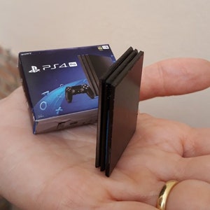 Puede incluir: Una réplica en miniatura de una consola PlayStation 4 Pro y su caja. La consola es negra y la caja es azul y negra con el logotipo de PlayStation y el texto "PS4 Pro".