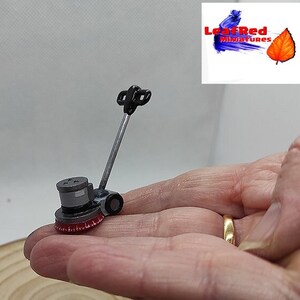 Puede incluir: Mini pulidora de suelo gris y negra con un cabezal de cepillo rojo. La pequeña herramienta de limpieza tiene un mango largo plateado. El logotipo de LeafRed Miniatures está en la esquina superior derecha. Sostenido en una mano.