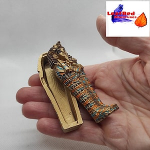 Puede incluir: Un sarcófago en miniatura pintado de oro con un diseño detallado, abierto para revelar una pequeña momia en miniatura pintada de oro en su interior. La momia está decorada con detalles azules y naranjas.