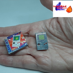 Puede incluir: Un Game Boy gris en miniatura con una pantalla azul y botones rojos, y una caja de juego Game Boy en miniatura con un Game Boy blanco y azul sobre un fondo colorido. Los artículos se sostienen en la mano de una persona.