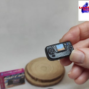 May include: A miniature black Sega Game Gear handheld video game console with a blue and white screen displaying a game. The console has a black and white button layout and is held in a hand.
