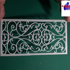 Può includere: Pannello decorativo in plastica bianca con un design dettagliato, intricato e simmetrico di motivi a spirale e a rotolo. Il pannello è su una superficie verde.