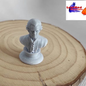 May include: A small, white, painted bust of a person with a detailed face and clothing. The bust is on a round, white base. The bust is on a wooden surface.