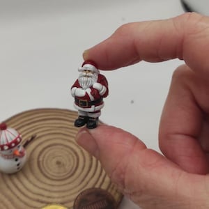 Puede incluir: Una figura en miniatura de Papá Noel con un traje rojo, barba y gorro blancos. La figura se sostiene entre los dedos, con un muñeco de nieve, un centavo y una moneda de 10 céntimos de fondo. La figura de Papá Noel mide aproximadamente 2,5 cm de alto.