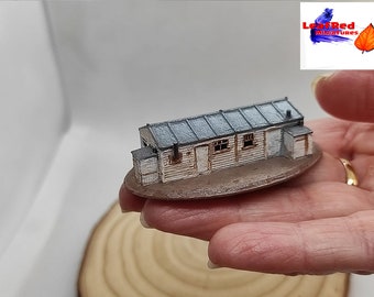 Puede incluir: Modelo en miniatura de un edificio de madera desgastada con tejado gris. La pequeña estructura presenta ventanas, puertas y una chimenea detalladas. El modelo está montado sobre una base ovalada marrón, mostrando una estética rústica.