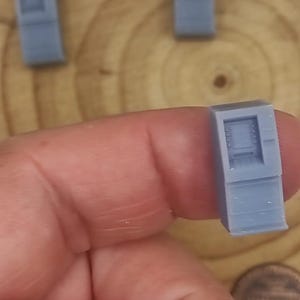 Puede incluir: Un modelo de computadora en miniatura de color azul claro se sostiene entre los dedos. El modelo tiene forma rectangular con una pantalla y un teclado. Otros dos modelos son visibles en el fondo. También se ve una moneda de un centavo.