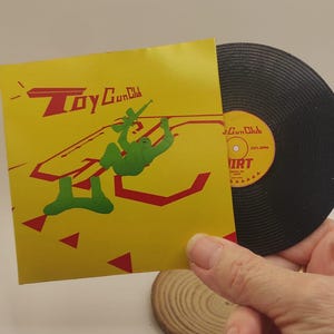 May include: A yellow square record sleeve featuring the red text Toy Gun Club and a green illustration of a figure holding a rifle. Beside it is a black vinyl record with a yellow center label displaying the text Gun Club and DIRT.