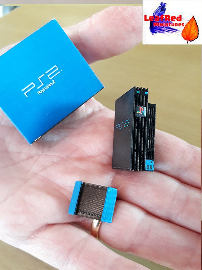 May include: A miniature model of a black PlayStation 2 console with a blue trim, a matching blue box with the text "PS2 PlayStation 2" and a small black and brown controller.