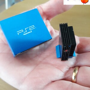 May include: A miniature replica of a PlayStation 2 console and its box. The console is black with a blue base and the box is blue with the PlayStation 2 logo and text.