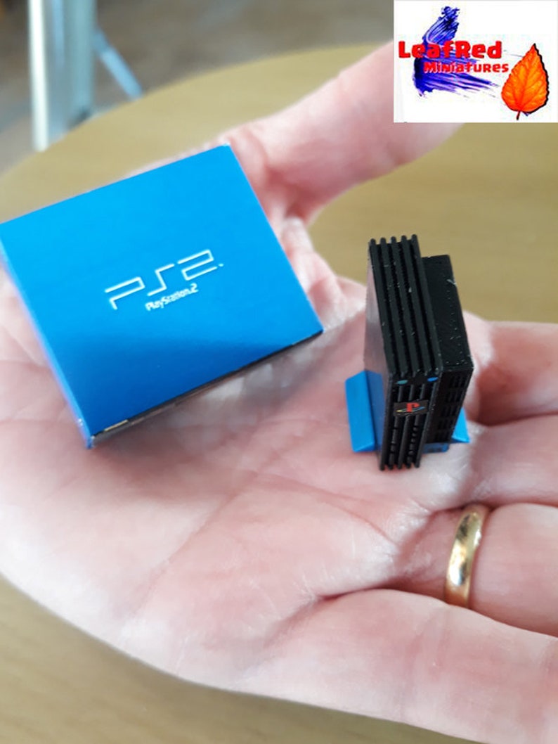 May include: A miniature replica of a PlayStation 2 console and its box. The console is black with a blue base and the box is blue with the PlayStation 2 logo and text.