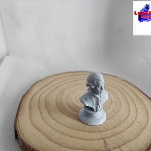 May include: A gray bust sculpture of a person with a hat on a wooden base. The bust is on a wooden surface.