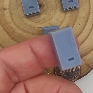 Puede incluir: Varios objetos pequeños, rectangulares, de color azul claro con un detalle rectangular empotrado. Los objetos se sostienen en una mano, con una moneda visible en el fondo. Parecen ser pequeñas piezas o componentes.
