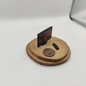 Op de afbeelding: Een miniatuurtelevisie met een zwart scherm en een rood en oranje beeld op het scherm. De televisie staat op een klein houten platform met een afstandsbediening en een cent.