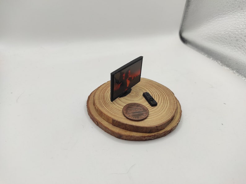 Op de afbeelding: Een miniatuurtelevisie met een zwart scherm en een rood en oranje beeld op het scherm. De televisie staat op een klein houten platform met een afstandsbediening en een cent.