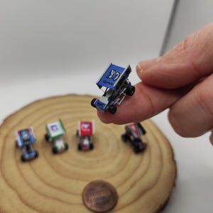 Puede incluir: Un coche de carreras miniatura azul con el número 97 impreso en el alerón lateral, sostenido entre los dedos. Cuatro coches de carreras miniatura adicionales en azul, verde, rojo y negro están dispuestos cerca, junto a una moneda de cobre.