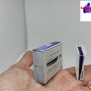 Puede incluir: Una consola PlayStation 5 en miniatura de color blanco y su caja. La caja es blanca con detalles azules y el texto "Play Like Never Before" en la parte delantera. La consola es blanca con una franja azul en el lateral.