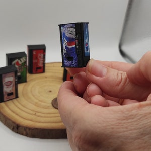 Puede incluir: Un conjunto de cinco máquinas expendedoras en miniatura, cada una con un diseño diferente. Las máquinas expendedoras son negras con imágenes coloridas de latas de refresco. Las máquinas expendedoras están sobre una base de madera.