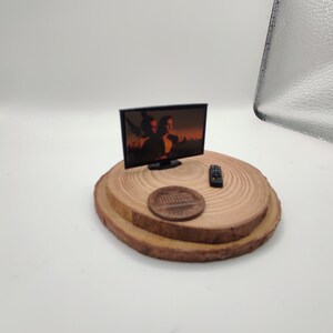 Op de afbeelding: Een miniatuur zwarte televisie met een scherm dat een sc&egrave;ne uit een film laat zien. De televisie staat op een houten platform met een afstandsbediening en een cent.