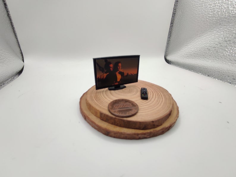 Op de afbeelding: Een miniatuur zwarte televisie met een scherm dat een sc&egrave;ne uit een film laat zien. De televisie staat op een houten platform met een afstandsbediening en een cent.