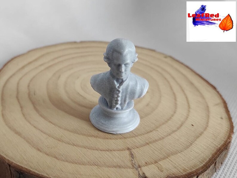 May include: A small, white, painted bust of a person with a detailed face and clothing. The bust is on a round, white base. The bust is on a wooden surface.
