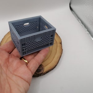 May include: A small, gray plastic crate with a grid pattern. The crate is empty and is sitting on a wooden surface.
