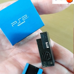 May include: A miniature blue PlayStation 2 box with the logo "PS2 PlayStation 2" on the front. Next to it is a miniature black PlayStation 2 console with a silver vent. There is also a miniature black controller with blue trim.