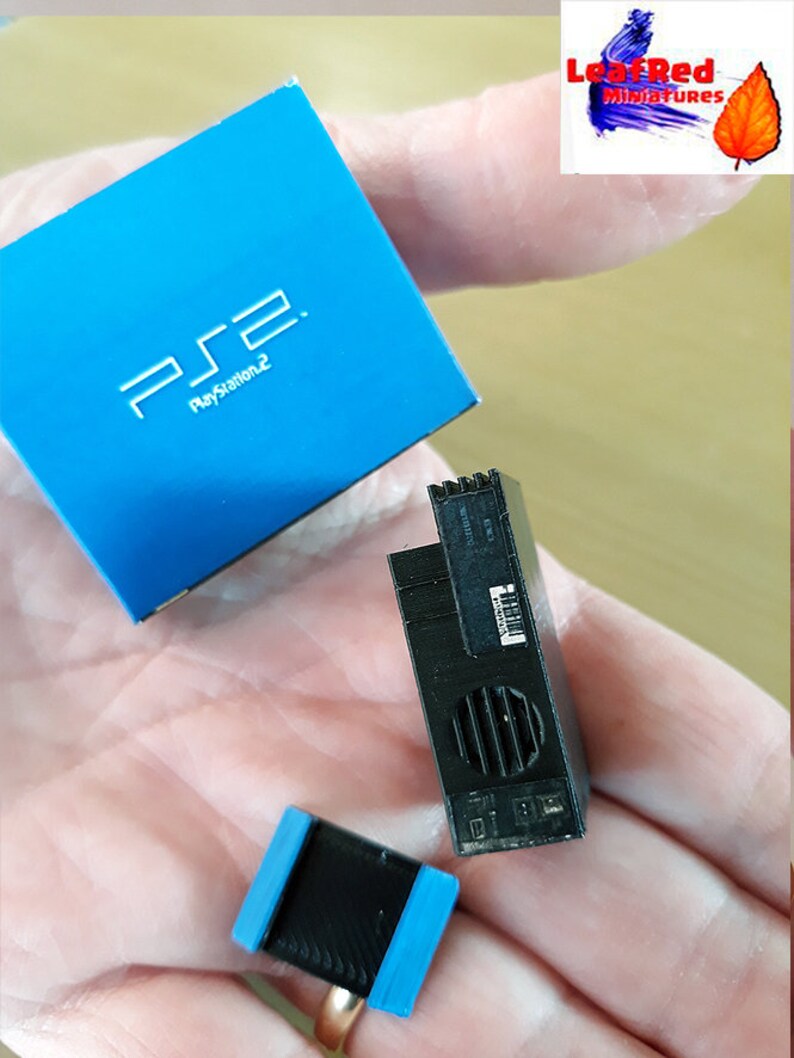 May include: A miniature blue PlayStation 2 box with the logo "PS2 PlayStation 2" on the front. Next to it is a miniature black PlayStation 2 console with a silver vent. There is also a miniature black controller with blue trim.