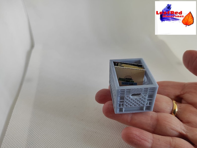 May include: A miniature gray plastic crate with a blue and white pattern, holding two miniature books. The crate is about 2 inches tall.
