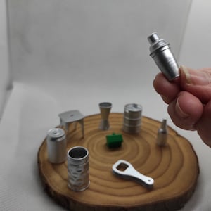 Könnte beinhalten: Ein Set an Miniatur-Barutensilien in Silber, darunter ein Shaker, eine Flasche, eine Dose, eine Tasse, ein Hocker, ein Fass, ein Flaschenöffner und ein grünes Plastikobjekt, alles auf einer Holzoberfläche angeordnet.