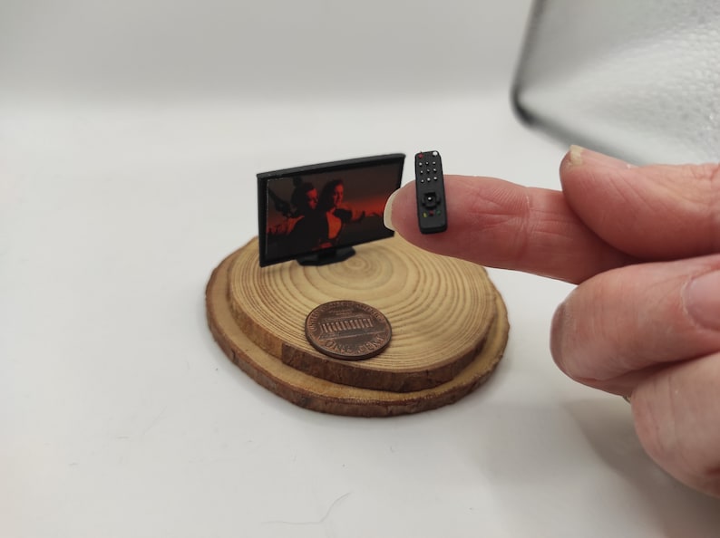 Op de afbeelding: Een miniatuurtelevisie met een zwart frame en een scherm dat een sc&egrave;ne uit een film laat zien. De televisie staat op een houten basis met een cent erop. Een miniatuur afstandsbediening wordt vastgehouden door een vinger.