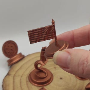 Puede incluir: Una bandera americana en miniatura sobre una base de madera, con un acabado de cobre. La bandera está izada en un pequeño mástil y está rodeada de otros objetos de color cobre sobre una superficie de madera.