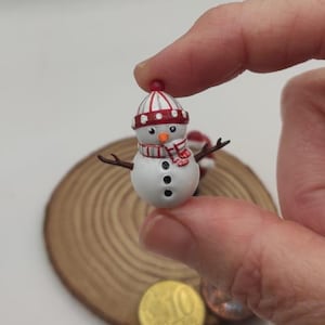 Könnte beinhalten: Eine Miniatur-Schneemannfigur mit weißem Körper, drei schwarzen Knöpfen, braunen zweigartigen Armen und einer orangefarbenen Karottennase. Der Schneemann trägt eine rot-weiß gestreifte Mütze mit passendem Schal und wird zwischen Fingern gehalten.