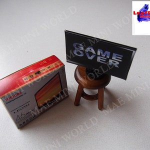 Op de afbeelding: Een miniatuur kartonnen doos voor een 46 inch Toshiba LED-tv met de tekst "Leading Innovation 170" en "LEDTV 46" op de doos. Een miniatuur zwart tv-scherm met de tekst "GAME OVER" erop staat op een kleine houten kruk.