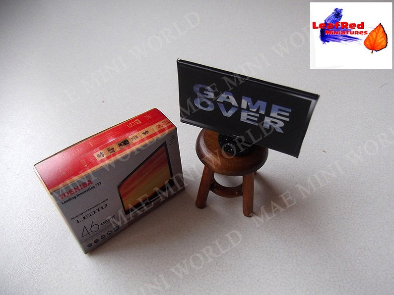 Op de afbeelding: Een miniatuur kartonnen doos voor een 46 inch Toshiba LED-tv met de tekst "Leading Innovation 170" en "LEDTV 46" op de doos. Een miniatuur zwart tv-scherm met de tekst "GAME OVER" erop staat op een kleine houten kruk.