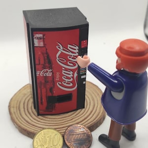 Könnte beinhalten: Eine Miniatur-Coca-Cola-Verkaufsmaschine in Rot und Schwarz mit dem Coca-Cola-Logo. Eine Spielfigur in einem blauen Mantel und rotem Hut greift nach der Maschine. Zwei Münzen, ein Penny und eine 10-Cent-Euro-Münze, liegen davor.