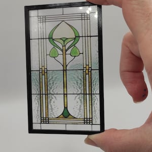 Puede incluir: Un panel de vitral con marco negro. El diseño presenta una planta verde estilizada con un centro blanco y detalles dorados.