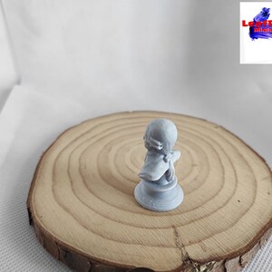 May include: A gray 3D printed miniature figurine of a person wearing a hat and a cloak. The figurine is standing on a wooden base.