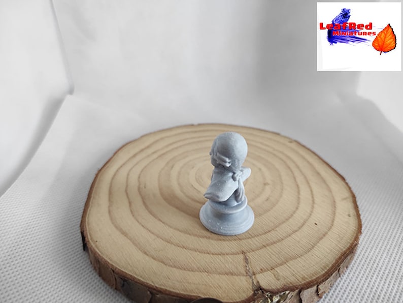 May include: A gray 3D printed miniature figurine of a person wearing a hat and a cloak. The figurine is standing on a wooden base.