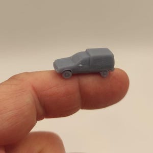 Puede incluir: Un pequeño camión de juguete gris con una zona de carga cubierta. El vehículo en miniatura tiene cuatro ruedas y descansa sobre un dedo. El camión es de color sólido y parece estar hecho de plástico o resina.