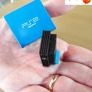 May include: A miniature black PlayStation 2 console with a blue base. The console is sitting on a hand with a gold ring. The console is next to a blue box with the PlayStation 2 logo.