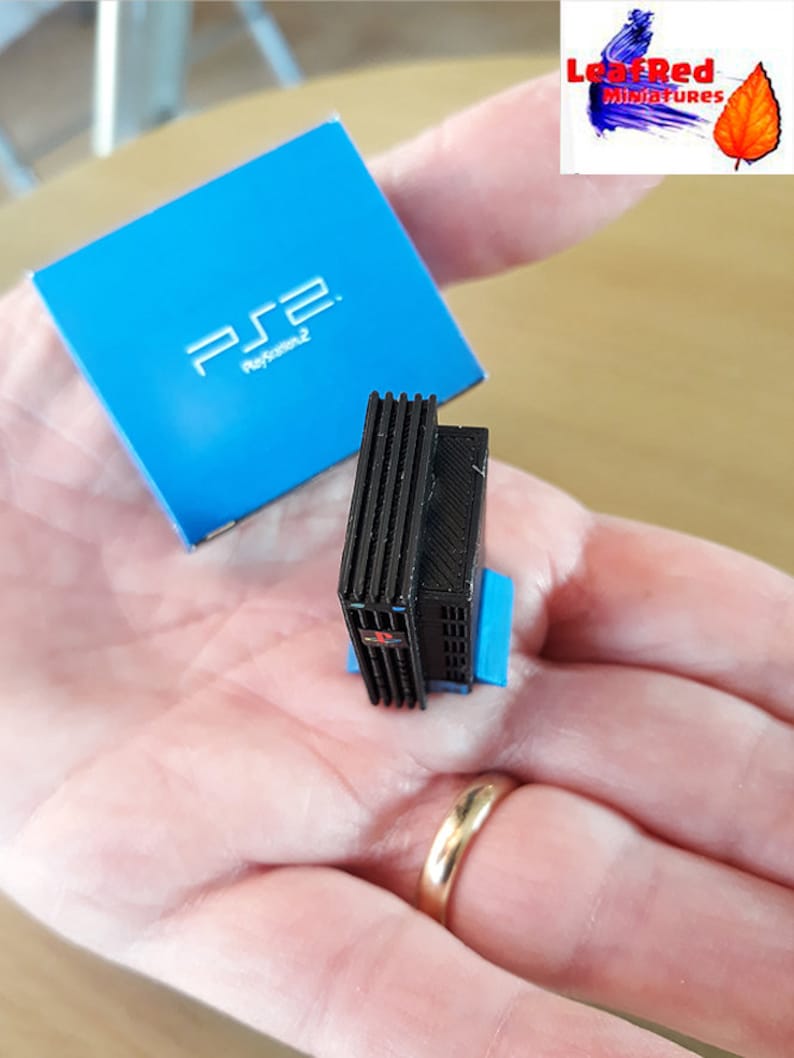 May include: A miniature black PlayStation 2 console with a blue base. The console is sitting on a hand with a gold ring. The console is next to a blue box with the PlayStation 2 logo.