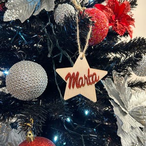 Puede incluir: Un árbol de Navidad negro decorado con adornos plateados y rojos, incluyendo un adorno en forma de estrella con el nombre "Marta" escrito en rojo.