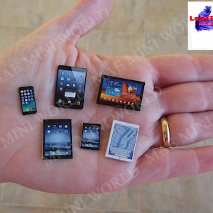 Puede incluir: Una mano sosteniendo cinco dispositivos electrónicos en miniatura, incluyendo un teléfono inteligente, una tableta y tres dispositivos más pequeños con pantallas. Los dispositivos son todos negros con pantallas coloridas.