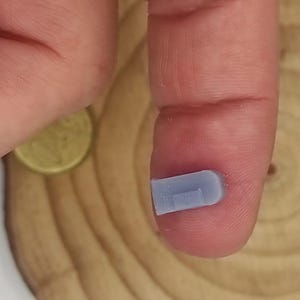 Puede incluir: Un pequeño objeto rectangular azul claro con un extremo curvo, sostenido entre un dedo y un pulgar. Una moneda de oro es visible en el fondo, junto con una superficie de madera con un patrón circular.