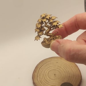 Puede incluir: Una figura de bonsái en miniatura de color dorado con un tronco curvado y grupos de hojas detallados, colocada en una pequeña maceta redonda. La escultura tiene un acabado metálico y se muestra a escala junto a una rodaja de madera y una moneda.
