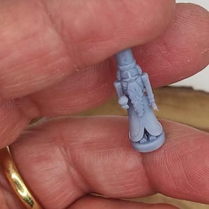 May include: A small, light blue figurine of a nutcracker soldier. The figurine has a detailed design, including a hat, beard, and uniform. The nutcracker is held between fingers, showcasing its size and intricate details.