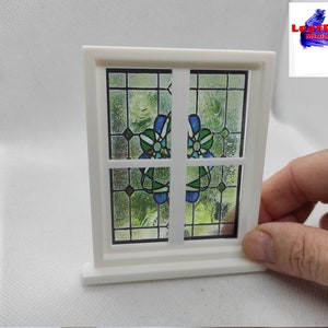 Puede incluir: Marco de ventana blanco en miniatura con un diseño de vitral que presenta un patrón floral azul y verde. La ventana está sobre una base blanca.