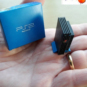 May include: A miniature black PlayStation 2 console with a blue base, next to a blue box with the PlayStation 2 logo. The console and box are on a person's hand. The image is labeled "LeafRed Miniatures".