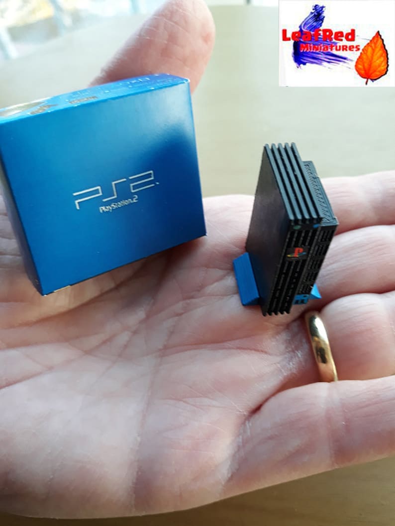 May include: A miniature black PlayStation 2 console with a blue base, next to a blue box with the PlayStation 2 logo. The console and box are on a person's hand. The image is labeled "LeafRed Miniatures".