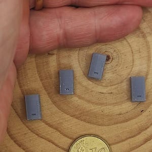 Puede incluir: Cuatro pequeños objetos rectangulares grises con un detalle de línea están dispuestos sobre una superficie de madera, junto a una moneda de 10 céntimos de euro. Los objetos parecen ser modelos o componentes en miniatura.